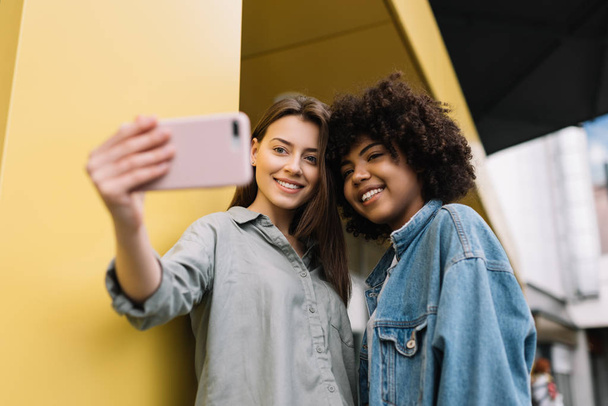 Positive hipsters friends holding mobile phone, taking pictures, laughing, having fun together indoors. Mixed race bloggers wearing stylish casual clothes streaming video online using smartphone and internet connection - 写真・画像