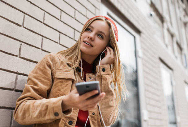 Attractive hipster woman holding mobile phone and  listening music outdoors. Music lover concept. Beautiful stylish female wearing casual clothes and red hat walking street, selects music in playlist. - 写真・画像