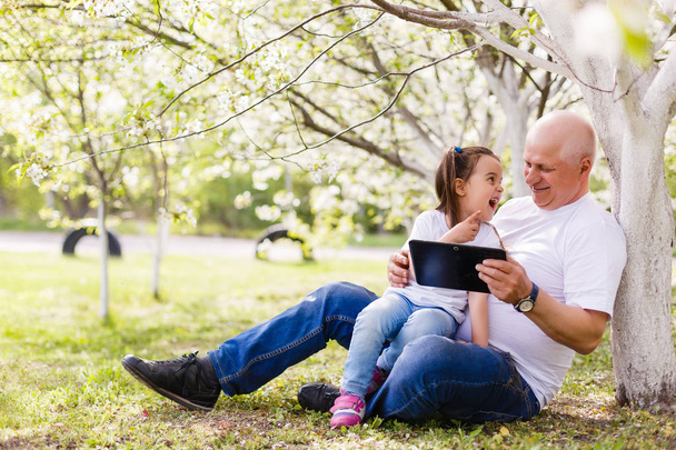Grandfather hugging granddaughter and using digital tablet sitting in park. - Photo, Image