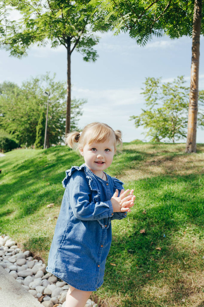 Pequeña niña de 3 años en un vestido azul vaqueros caminando en un parque en un día cálido y soleado
 - Foto, Imagen