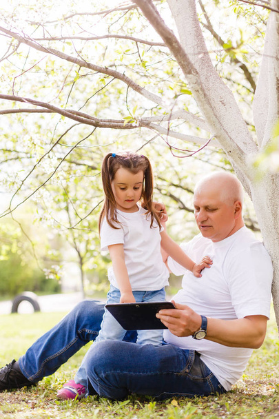 Dziadek wnuczka przytulanie i za pomocą cyfrowego tabletu siedzi w parku. - Zdjęcie, obraz