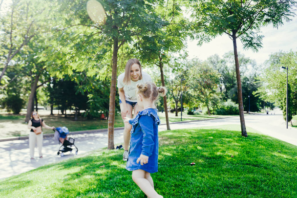 Portrait of 3 years old girl running away from her mother while playing on a lawn in a city park on a warm sunny day. Nanny with pram on the background - Фото, изображение