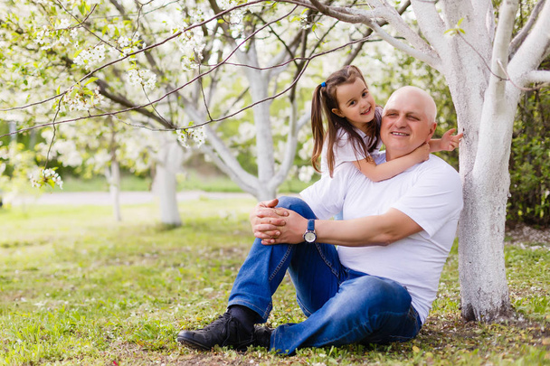 少女庭園で桜開花ツリーの下に座っている彼女の祖父を抱き締める - 写真・画像