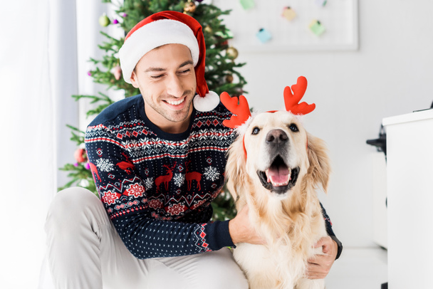man in christmas sweater and santa hat stroking golden retriever in deer horns  - Photo, Image