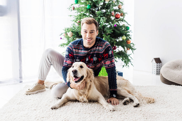 happy man in sweater with golden retriever dog sitting near christmas tree - Фото, изображение