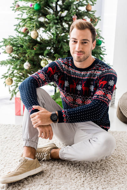 handsome man in winter sweater sitting near christmas tree  - Fotó, kép