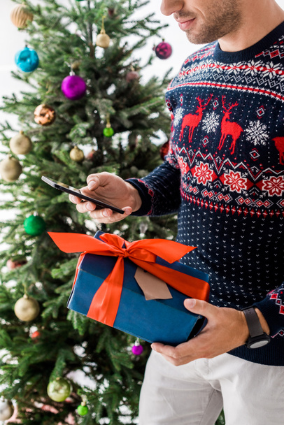 cropped view of man in christmas sweater with gift box and smartphone - Foto, immagini