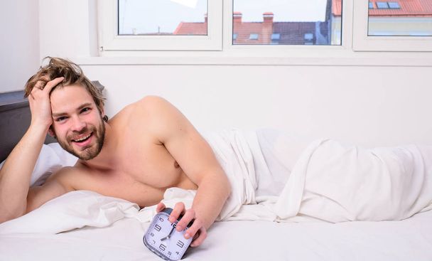 Stick schedule same bedtime wake up time. Enough sleep for him. Regulate your bodys clock. Man unshaven tousled hair wakeful face having rest. Good morning. Man unshaven lay bed hold alarm clock - Фото, зображення
