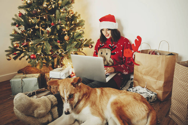 happy girl in santa hat working on laptop and sitting with cute dog at golden beautiful christmas tree with lights and presents in festive room. family atmospheric   moments. freelance - Foto, Bild