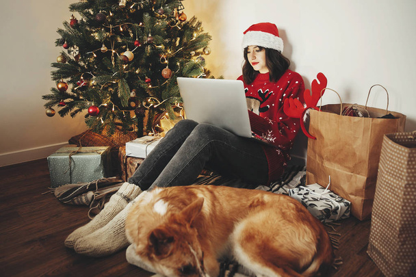 Menina feliz em santa chapéu de compras on-line no laptop e sentado com o cão bonito na árvore de Natal bonita dourada com luzes e presentes na sala festiva. venda de Natal e conceito de desconto - Foto, Imagem