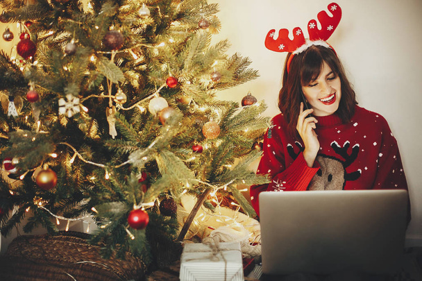 όμορφο κορίτσι στο τάρανδος ελαφοκέρατο μιλάμε στο τηλέφωνο και κάθεστε με το laptop στην Χρυσή χριστουγεννιάτικο δέντρο με φώτα και παρουσιάζει στην εορταστική αίθουσα. Χριστούγεννα αγορές online έννοια - Φωτογραφία, εικόνα