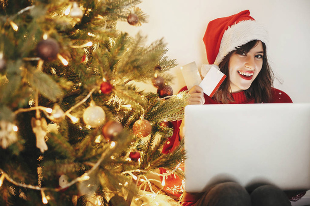 hermosa mujer joven en sombrero de santa celebración de tarjetas de crédito y sentarse con el ordenador portátil en el hermoso árbol de Navidad de oro con luces y regalos en la habitación festiva. compras y ventas de Navidad
 - Foto, Imagen