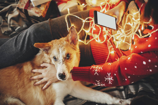 Mädchen hält Telefon mit leerem Bildschirm und umarmt Hund unter goldenen schönen Weihnachtsbaum mit Lichtern und Geschenken in festlichem Raum. Winterliche atmosphärische Momente. frohe Feiertage - Foto, Bild