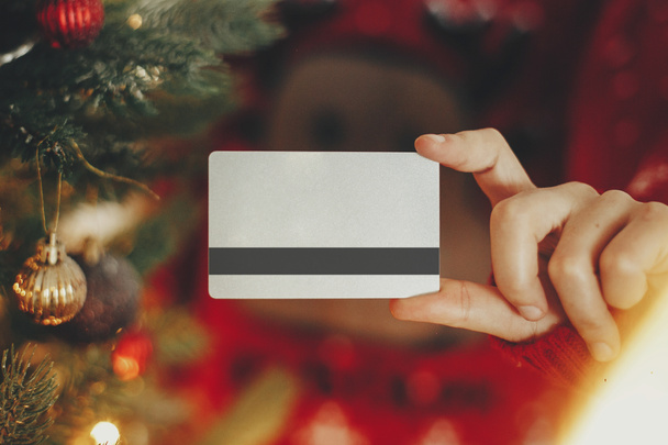 χέρι που κρατά γκρο πλαν πιστωτική κάρτα σε φόντο χρυσό όμορφο χριστουγεννιάτικο δέντρο με φώτα στην εορταστική αίθουσα. Χριστουγεννιάτικα ψώνια και πώληση έννοια. έκπτωση. Μαύρη Παρασκευή - Φωτογραφία, εικόνα