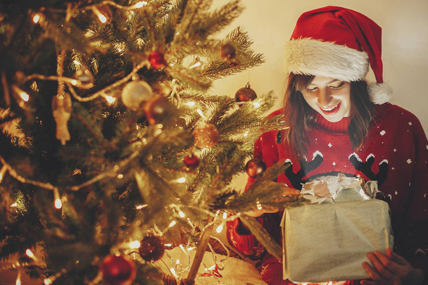 glückliches Mädchen mit Weihnachtsmütze öffnet magische Weihnachtsgeschenkbox an goldenen schönen Weihnachtsbaum mit Lichtern und Geschenken in festlichem Raum. Winterliche atmosphärische Momente. frohe Feiertage - Foto, Bild