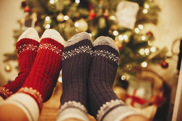 stylish festive socks on couple legs on background of golden beautiful christmas tree with lights in festive room. family relax time. cozy winter holidays. warm atmospheric moment - Photo, Image