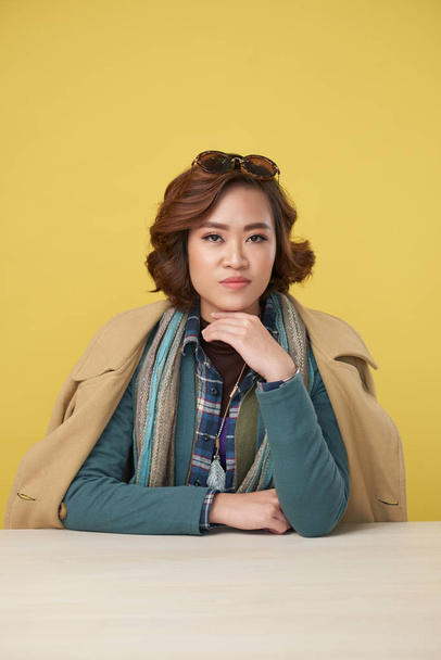 Portrait of confident Asian woman wearing autumn clothes sitting at table on yellow background - Foto, Imagem