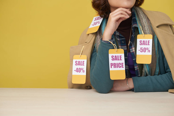 Mujer irreconocible que usa capas de ropa con etiquetas de venta mientras está sentada en la mesa sobre fondo amarillo
 - Foto, Imagen