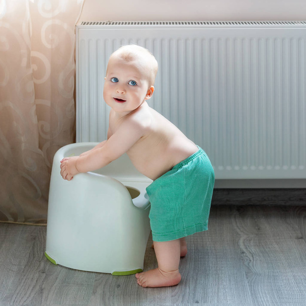 Cute little blond caucasian boy in funny green shorts playing with white potty indoor. Adorable child learning about potty-chair during game. Smiling positive kid with heating radiator on background. - Photo, Image