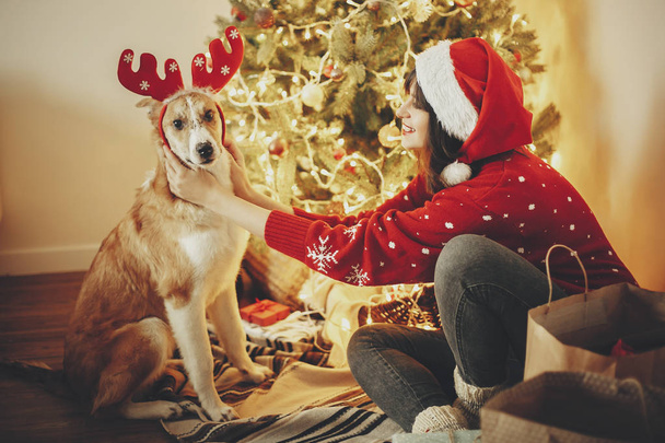 a lány aranyos kutya rénszarvas agancs, háttérben a golden gyönyörű karácsonyfa fények ünnepi szobában elhelyezés. kutyus imádnivaló szeme: ragyogó megvilágítás. családi téli ünnepek - Fotó, kép