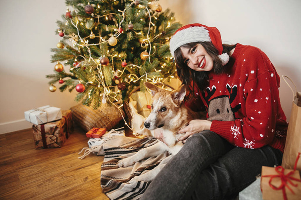 menina feliz em santa chapéu abraçando com cão bonito no fundo da bela árvore de Natal dourada com luzes na sala festiva. família momentos atmosféricos quentes. férias de inverno
 - Foto, Imagem