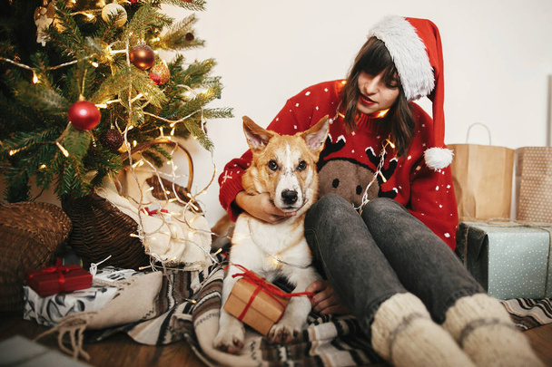 glückliches Mädchen mit Weihnachtsmütze umarmt mit niedlichem Hund auf dem Hintergrund der goldenen schönen Weihnachtsbaum mit Lichtern und Geschenken und Geschenke in festlichem Raum. Warme Momente in der Familie - Foto, Bild
