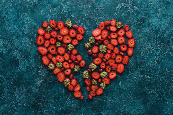 bovenaanzicht van gehalveerde hart teken gemaakt van rijpe aardbeien op blauwe beton oppervlak - Foto, afbeelding