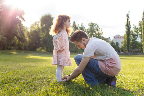 A father helping his little daughter with her shoes in the park. Curly kid girl in beautiful dress - Foto, Imagen