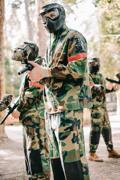 male paintball player in uniform and goggle mask looking at camera and standing with his team outdoors - Photo, Image