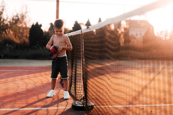 Μικρό αγόρι στέκεται στην παιδική χαρά του τένις και βάζοντας γάντια στα χέρια του. Προετοιμασία τον εαυτό για την εξωτερική εκπαίδευση. - Φωτογραφία, εικόνα