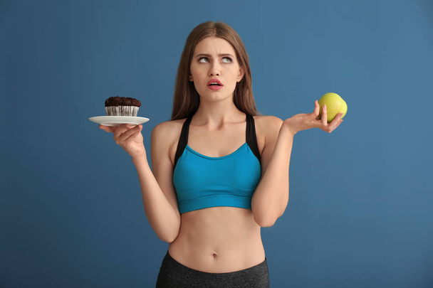 Молода жінка в спортивному одязі вибирає між тортами та яблуком на кольоровому фоні
 - Фото, зображення