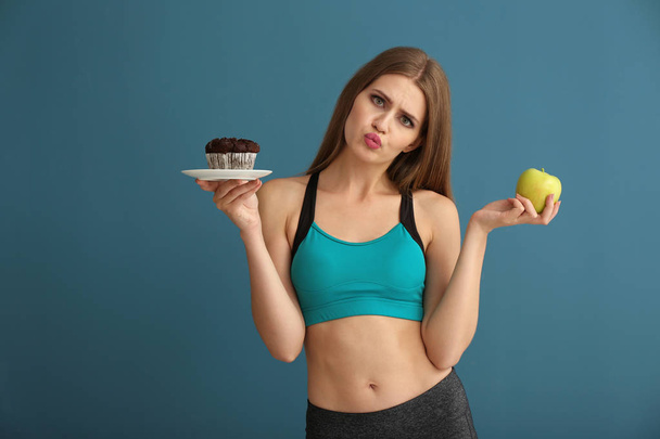 Jonge vrouw in sportkleding kiezen tussen taart en apple op kleur achtergrond - Foto, afbeelding