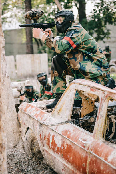 male paintball player aiming by marker gun and sitting on broken car while his team standing behind outdoors  - Photo, Image