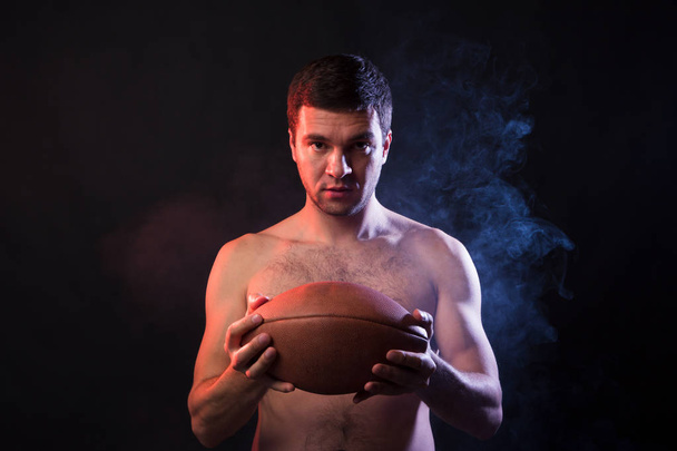プロのラグビーのスポーツマンは、彼の手でラグビー ボールを保持します。煙の黒い背景にスタジオ ポートレート。大学間の競争の勝者の概念 - 写真・画像