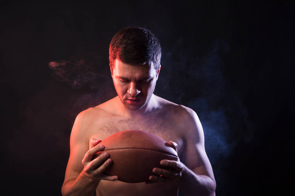 Profesionální ragby sportovec drží rugby míč v jeho rukou. Studiový portrét na černém pozadí v kouři. Koncept vítězem soutěží mezi vysoké školy - Fotografie, Obrázek
