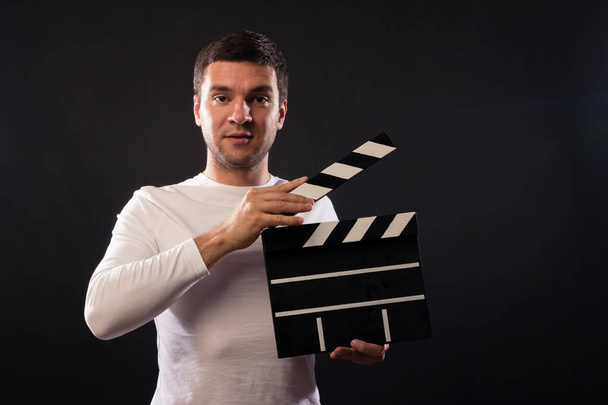 Young man of Caucasian appearance is holding a clapperboard. Portrait shot in studio on a black background with smoke and red and blue studio light.  - Photo, Image