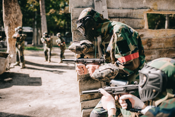 selective focus of paintball player in protective mask holding marker gun and his teammate hiding behind wooden wall outdoors - Photo, Image