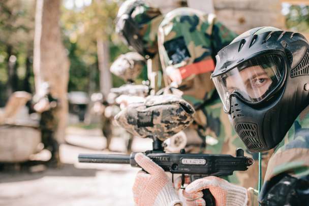 portrait of male paintball player in protective mask holding marker gun and looking at camera outdoors  - Photo, Image