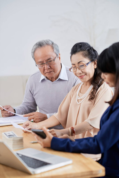 senior Asian couple sitting at modern office and paying for their new apartment to pretty realtor after successful completion of meeting - Photo, Image