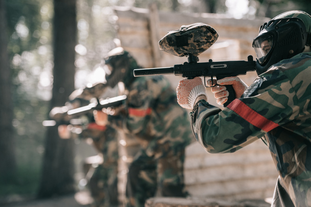 selective focus of paintball player in goggle mask aiming with marker gun and his team on background outdoors - Photo, Image
