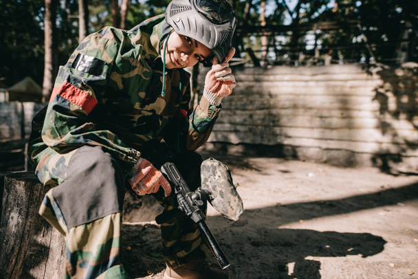glimlachend jonge vrouwelijke paintballer in camouflage uniforme zitten met paintball pistool buitenshuis  - Foto, afbeelding