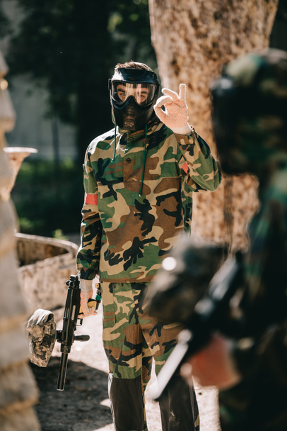 männlicher Paintballspieler in Camouflage und Brillenmaske, der seinem Team im Freien ein OK-Zeichen zeigt - Foto, Bild
