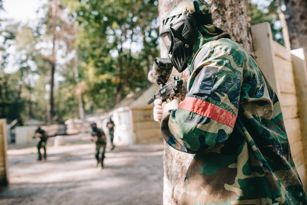 male paintballer in camouflage and protective mask holding marker gun and hiding behind tree while other team running behind outdoors  - Foto, afbeelding