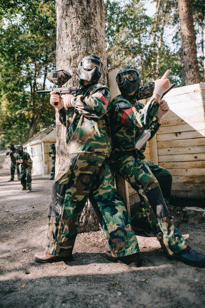 male paintball player in goggle mask and camouflage holding marker gun and pointing by finger to teammate near tree outdoors  - Photo, Image