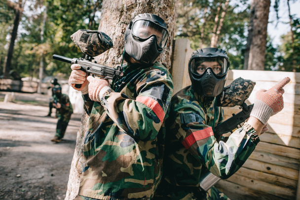 male paintball player in camouflage and goggle mask pointing by finger to his teammate with marker gun standing near tree outdoors - Photo, Image