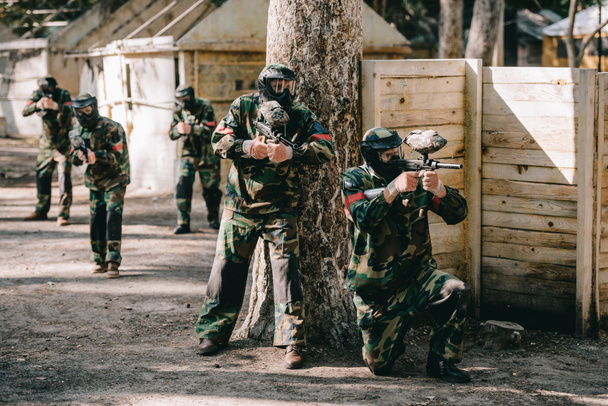 paintball players wearing camouflage and goggle masks shooting by markers guns near tree and their team on background outdoors - Photo, Image
