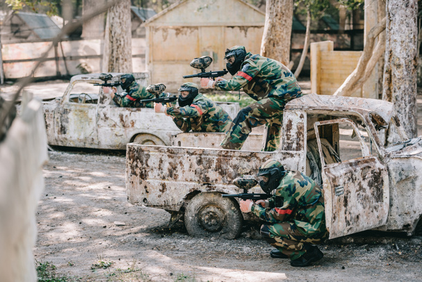 paintball team in camouflage and goggle masks aiming with marker guns from broken cars outdoors - Photo, Image