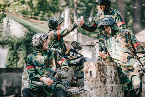 happy paintball team in camouflage celebrating victory and shaking hands on broken car outdoors  - Photo, Image