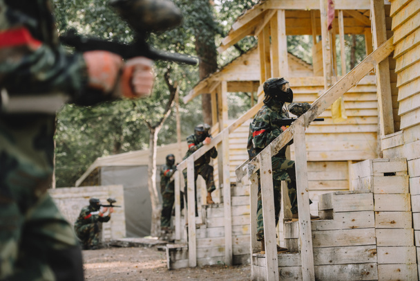 paintball team in uniform and protective masks standing on staircase of wooden towers with paintball guns outdoors - Photo, Image