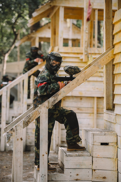 female paintballer in camouflage and goggle mask with marker gun standing on staircase of wooden tower outdoors - Photo, Image